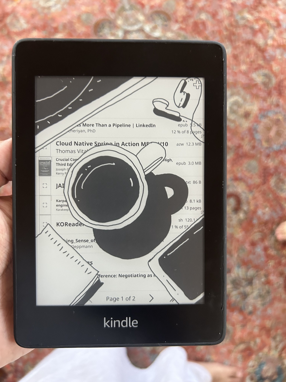 A Kindle Paperwhite running KOReader with a custom coffee-cup screensaver over its library